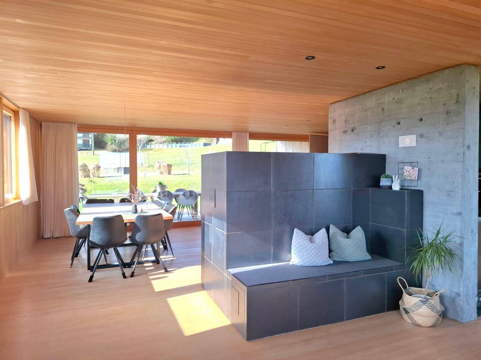 Offener Wohn-Essbereich mit Holzboden, Kachelofen und großen Fenstern mit Gartenblick.