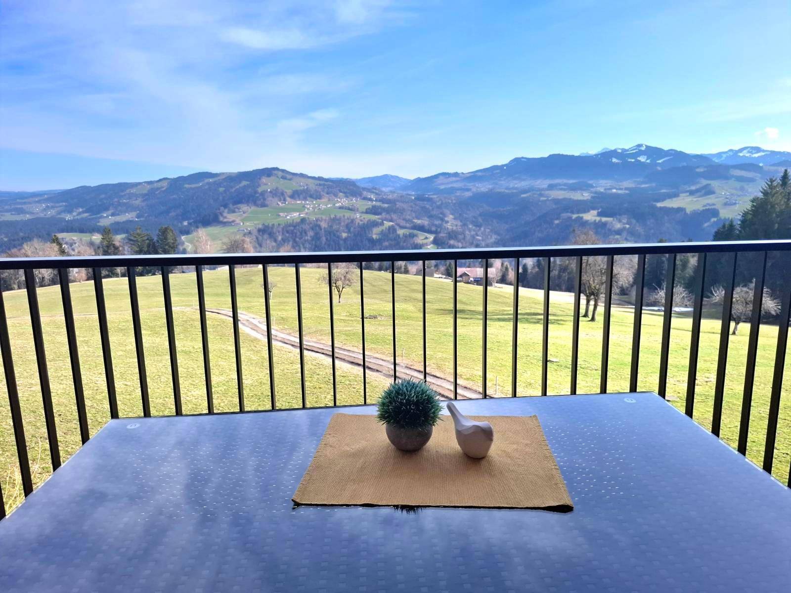 Balkon mit Tisch und Blick auf die weite Berglandschaft und grüne Hügel unter blauem Himmel.