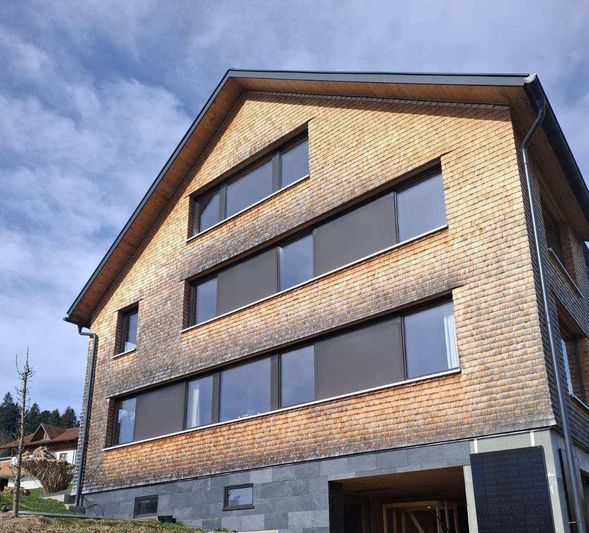 Detailansicht der Holzschindelfassade mit modernen Fenstern und Dachüberstand unter bewölktem Himmel.