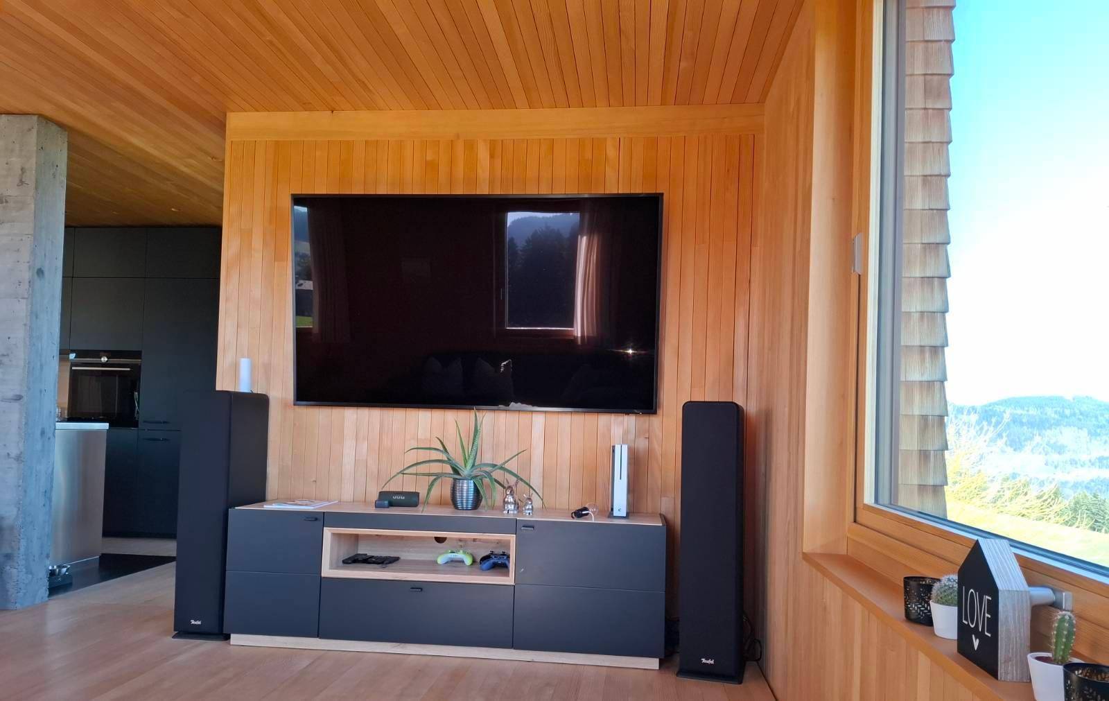 Wohnzimmer mit Holzwänden, großem Fernseher und Blick auf die Natur durch ein großes Fenster.