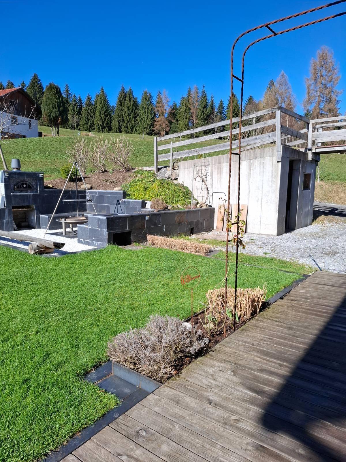 Gartenbereich mit Grillplatz und Rasenfläche, umgeben von Bäumen und einem Holzzaun.