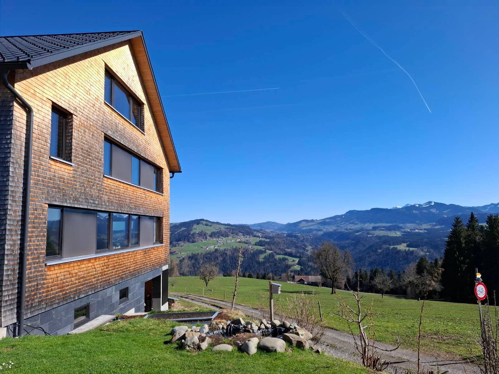 Modernes Holzhaus mit Schindelfassade und weitem Blick auf die Berge und grüne Hügel unter blauem Himmel.