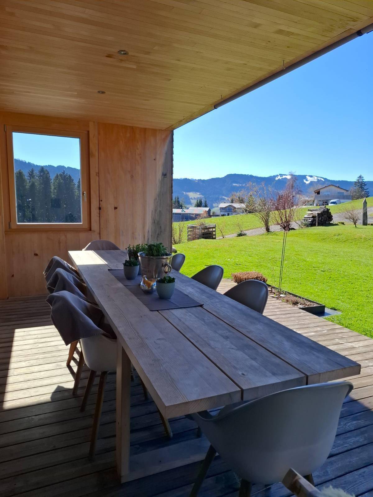 Überdachte Terrasse mit großem Holztisch und Stühlen, umgeben von grüner Landschaft und Bergblick.