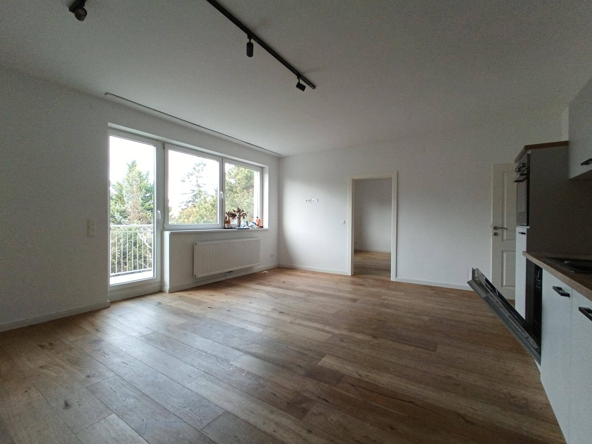 Lichtdurchfluteter Wohnbereich mit Holzboden, großen Fenstern und Zugang zum Balkon.