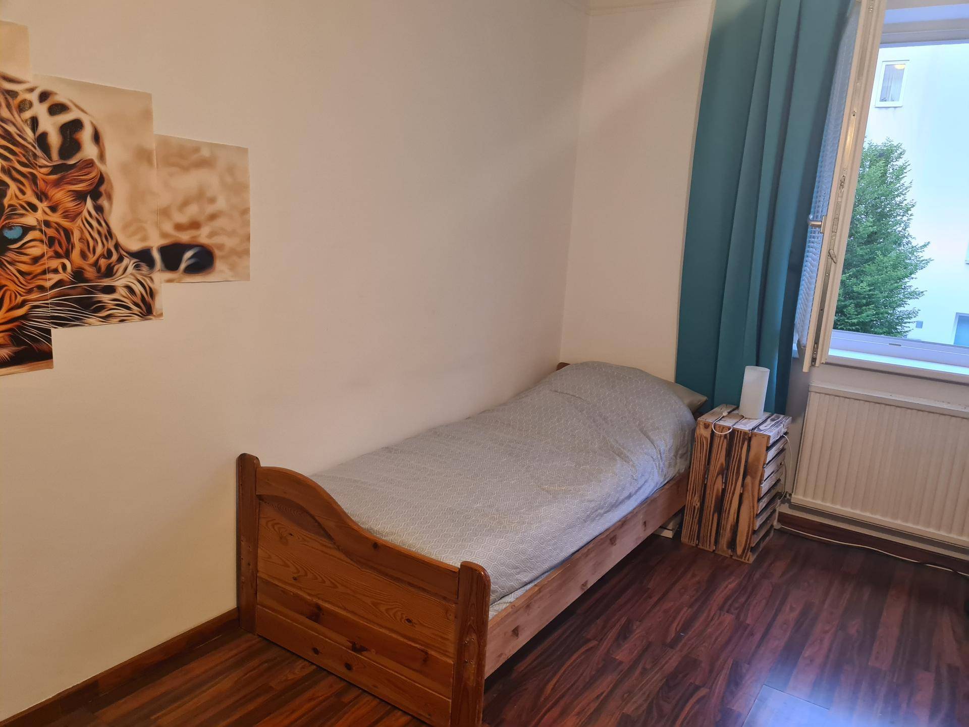 Gemütliches Schlafzimmer mit Einzelbett, Holzboden und einem Fenster mit blauem Vorhang.