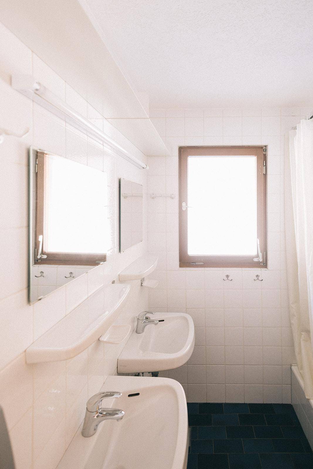 Helles Badezimmer mit zwei Waschbecken, weißen Fliesen und Fenster, einfach und funktional.