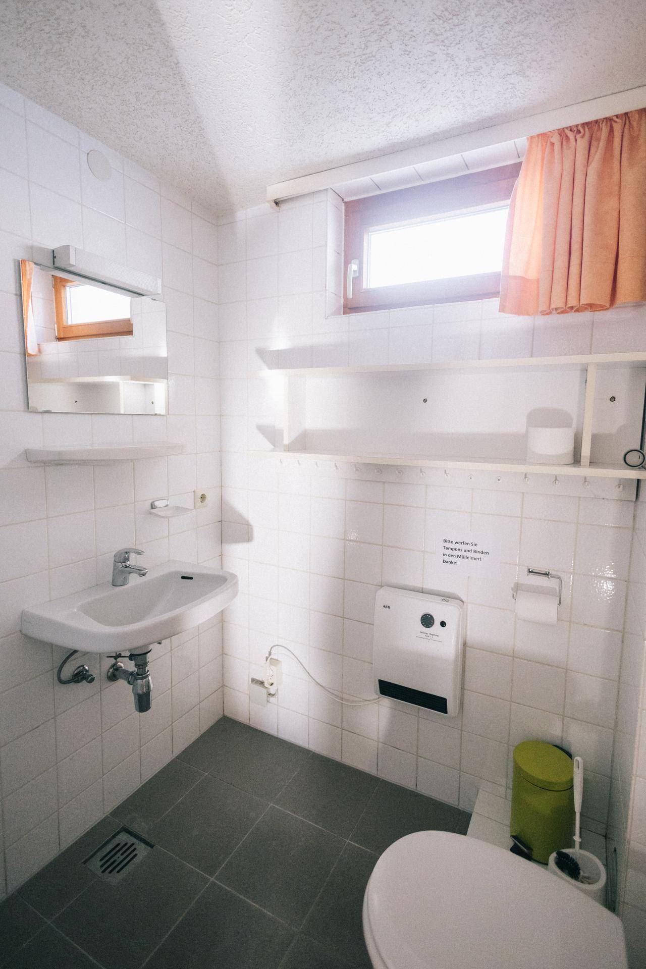 Badezimmer mit WC, Waschbecken und Fenster, komplett gefliest in Weiß und Grau, einfach gehalten.