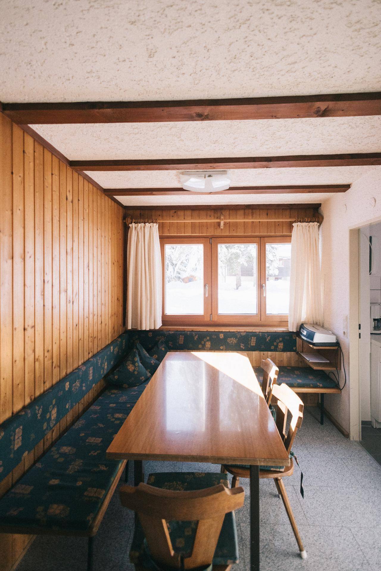 Heller Wohn-Essbereich mit Holztisch, Sitzbank und Blick ins Grüne durch große Fenster, mit Holzbalken.