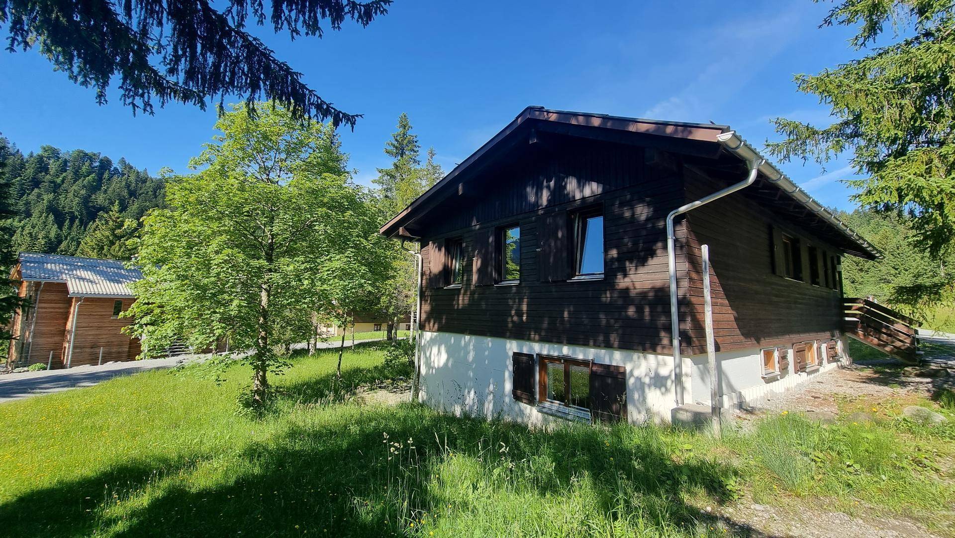 Charmantes Holzhaus in ländlicher Umgebung mit viel Grün und Bergblick unter blauem Himmel.