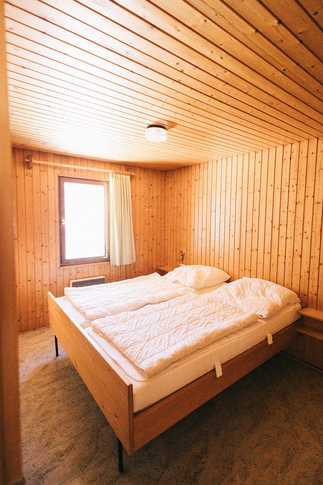 Gemütliches Schlafzimmer mit Doppelbett und Holzvertäfelung, hell durch das Fenster, einfach eingerichtet.