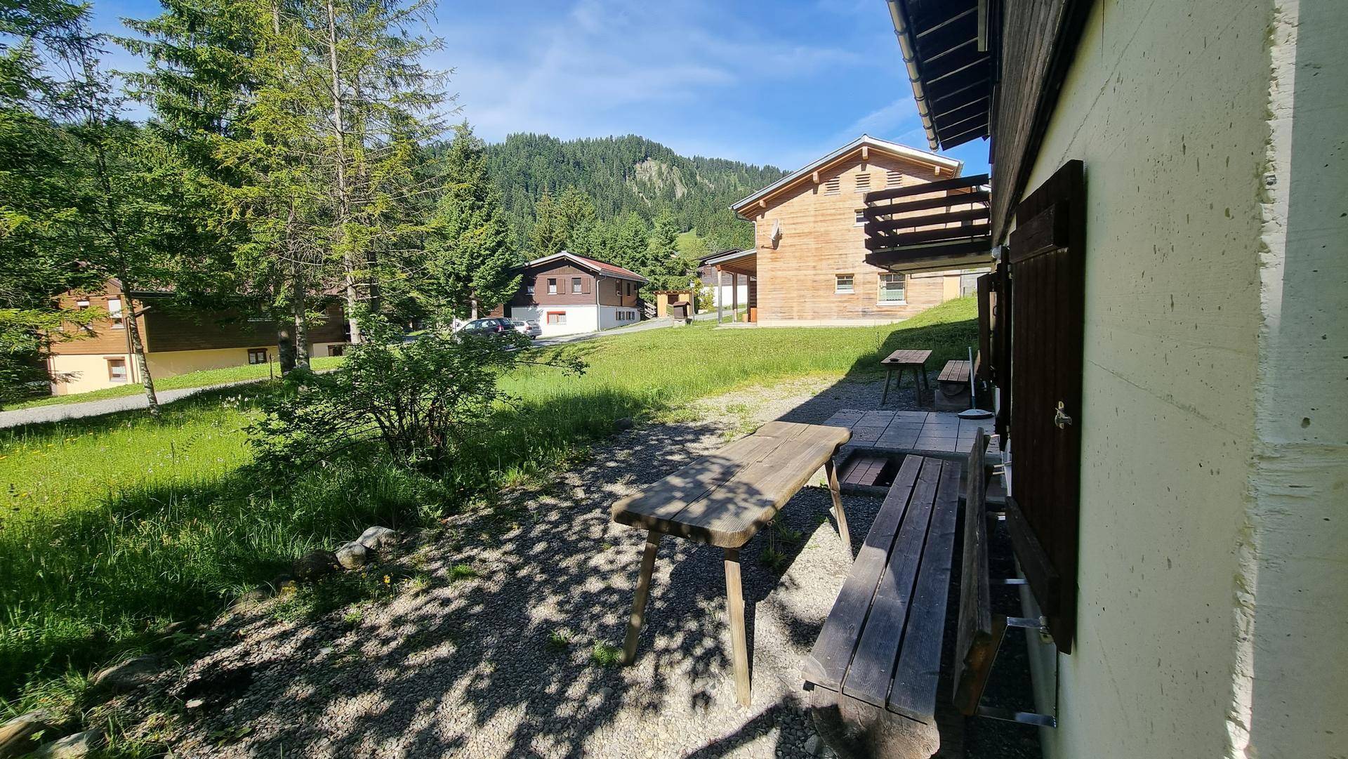 Außenbereich mit Holztisch und Bänken, umgeben von grüner Wiese und Bäumen im Sonnenschein.