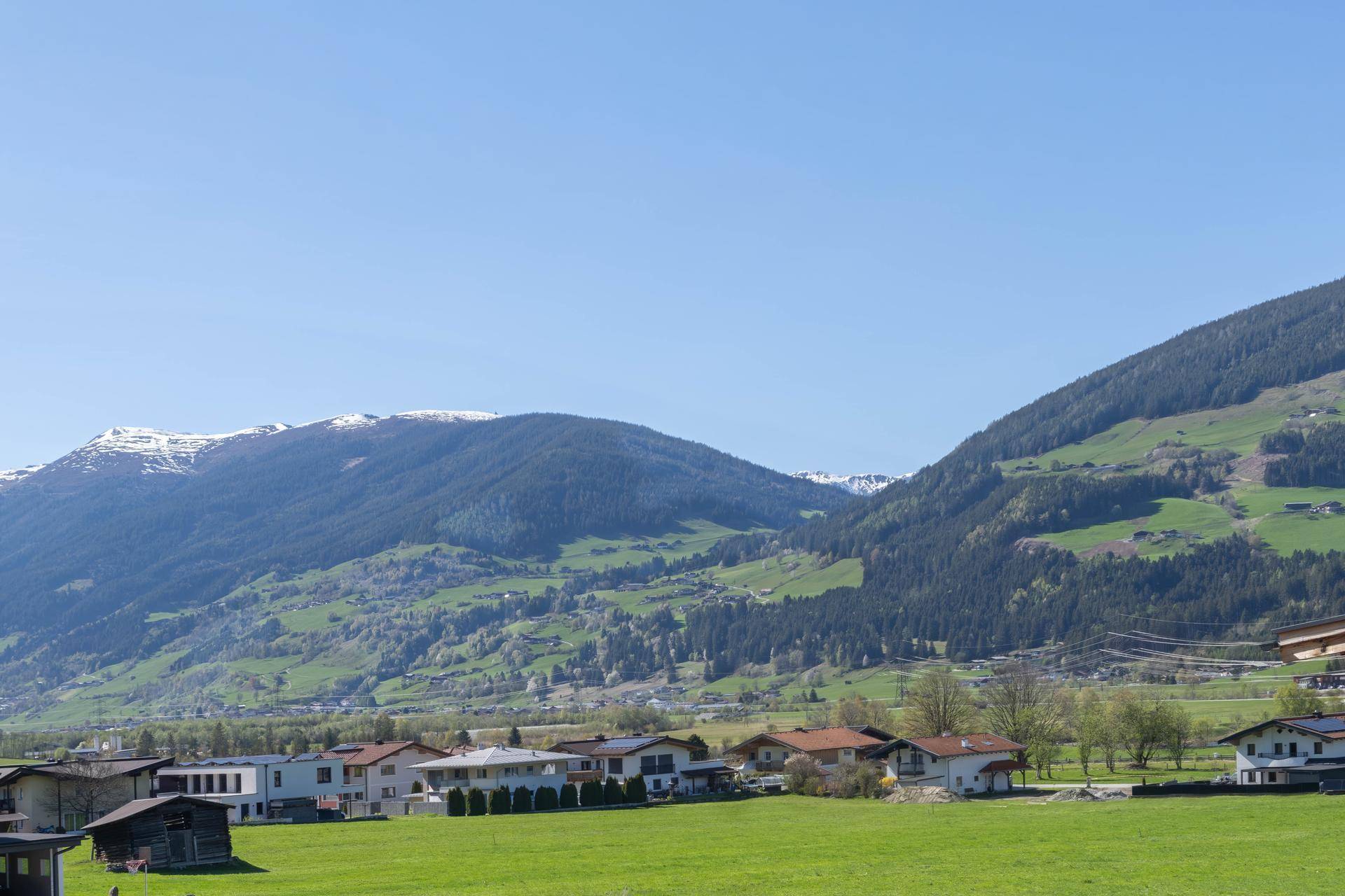 Weitläufige grüne Landschaft mit einem Dorf im Tal und schneebedeckten Bergen im Hintergrund unter blauem Himmel.