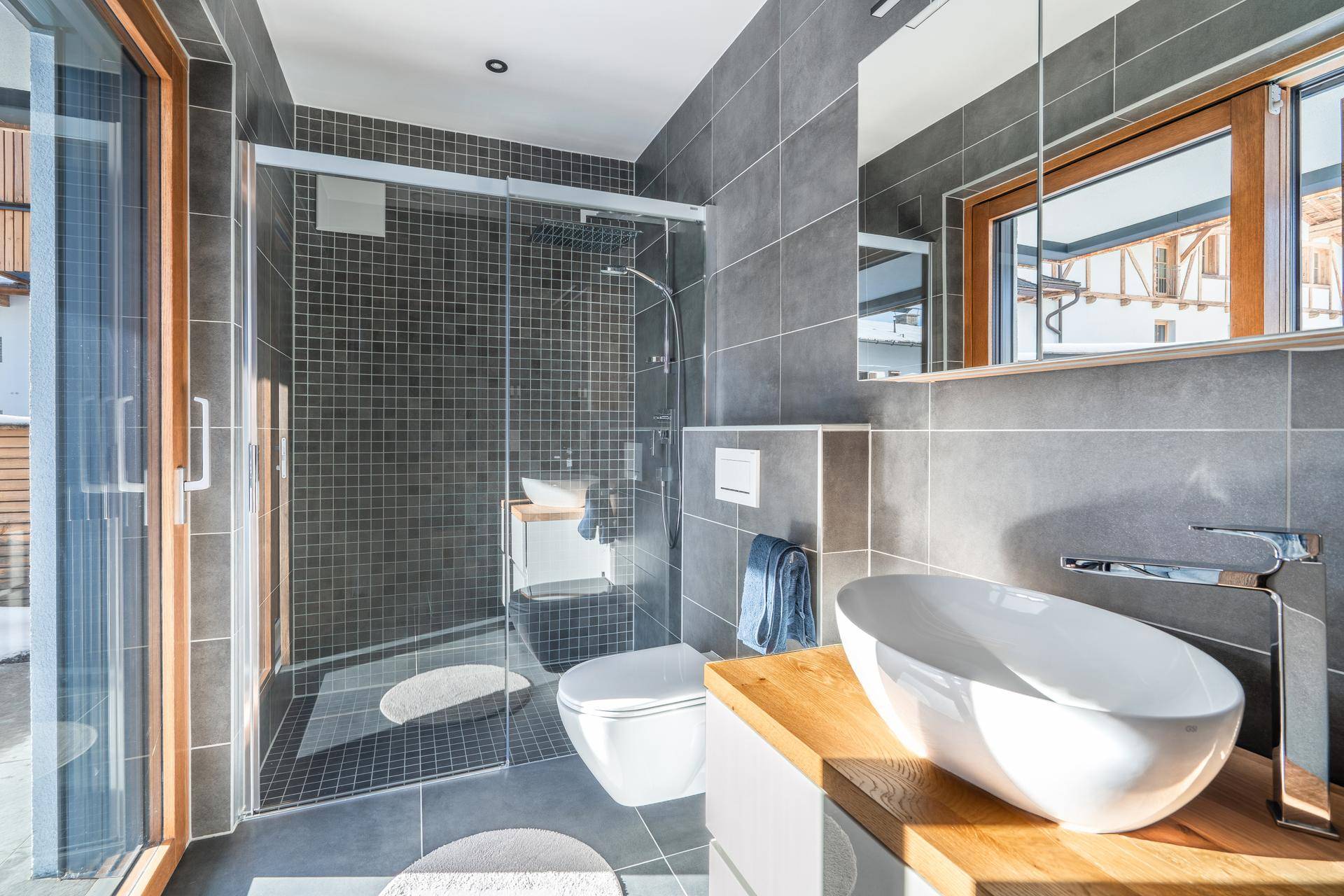 Modernes Badezimmer mit dunkelgrauen Fliesen, ebenerdiger Dusche und stilvollem Waschbecken auf Holzplatte.