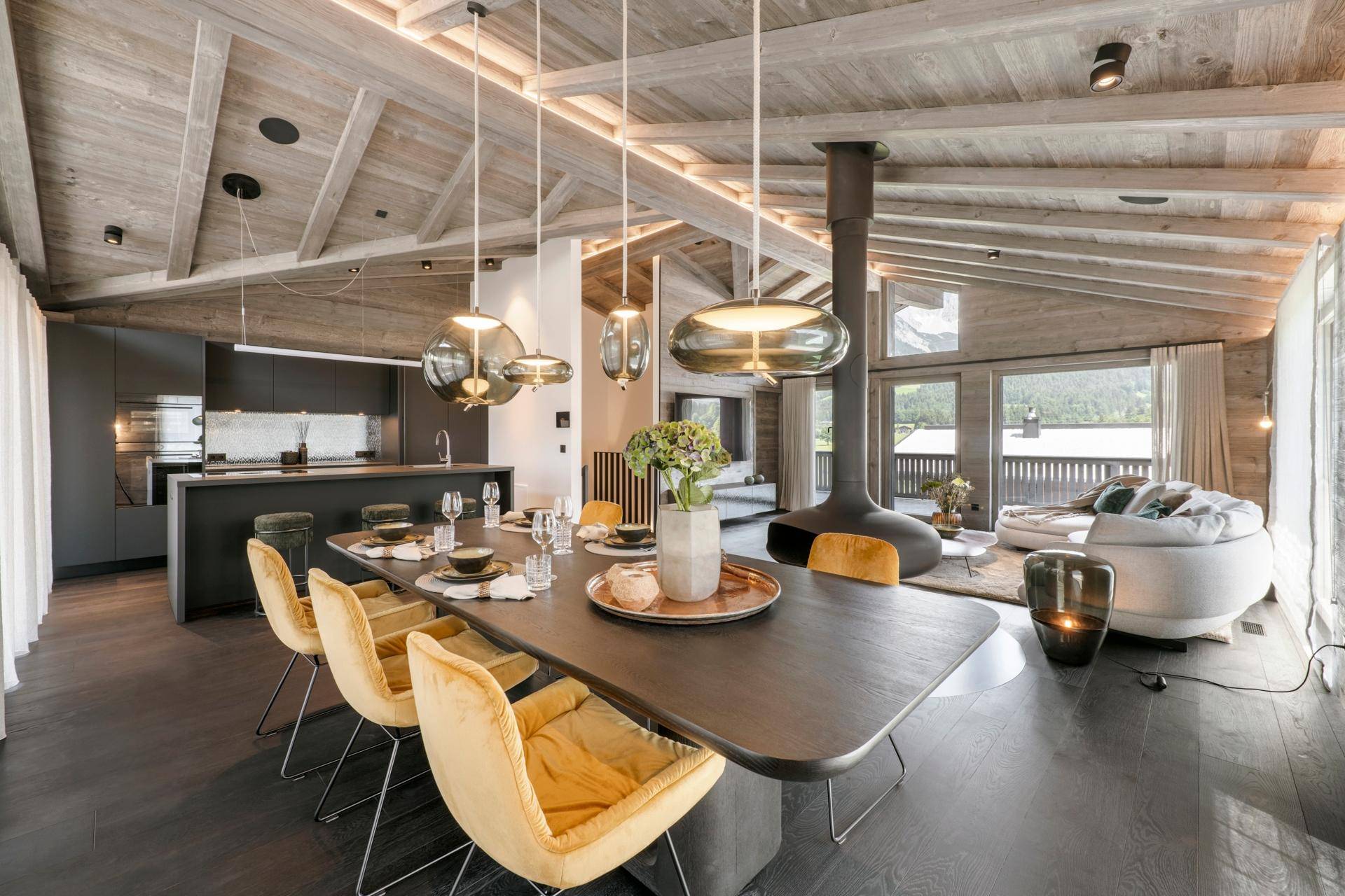 Moderner Essbereich mit Designerleuchten, Holzbalkendecke und offener Küche im Hintergrund.
