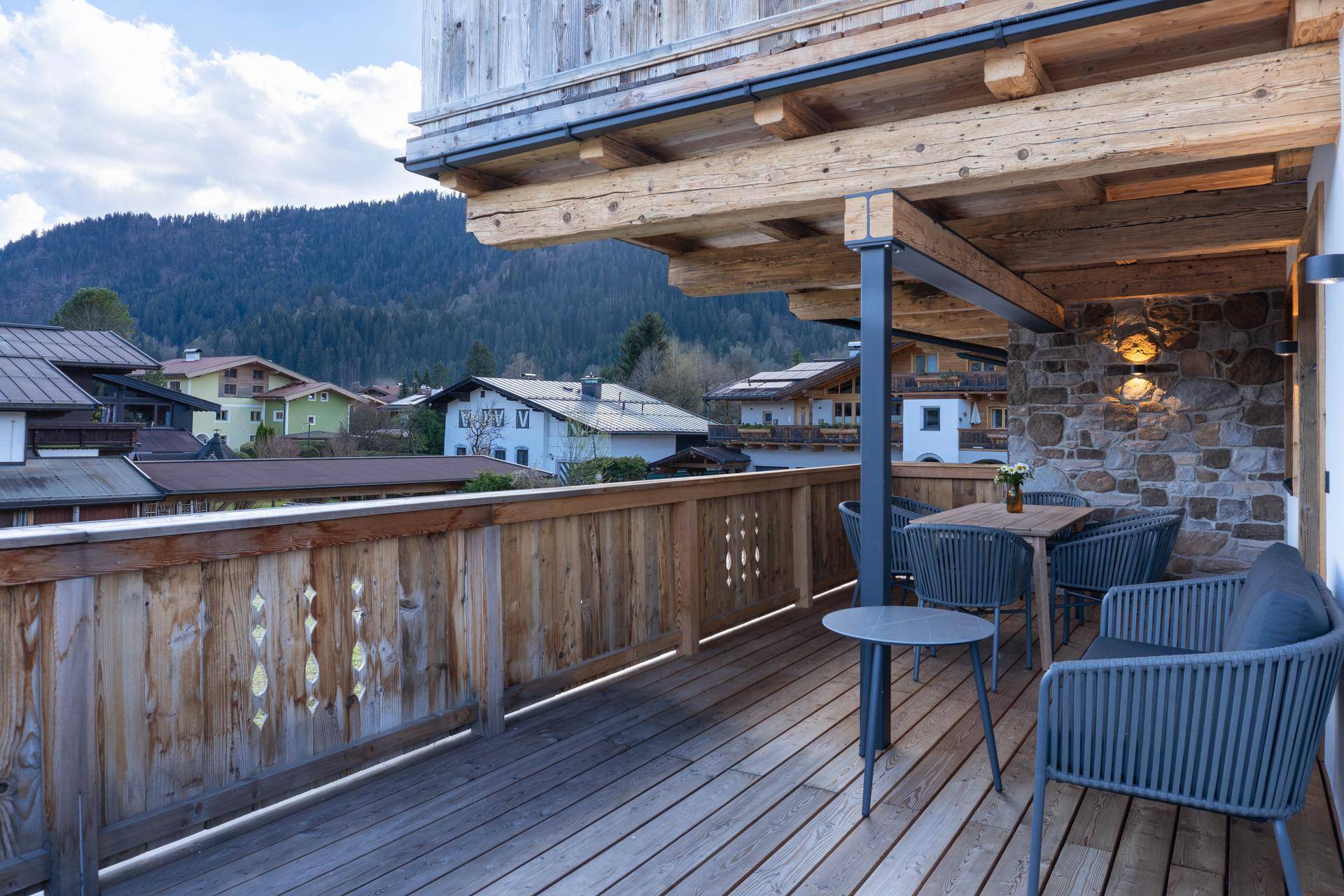 Möblierter Holzbalkon mit Steinwand, Sitzgelegenheiten und Blick auf die umliegende Landschaft.