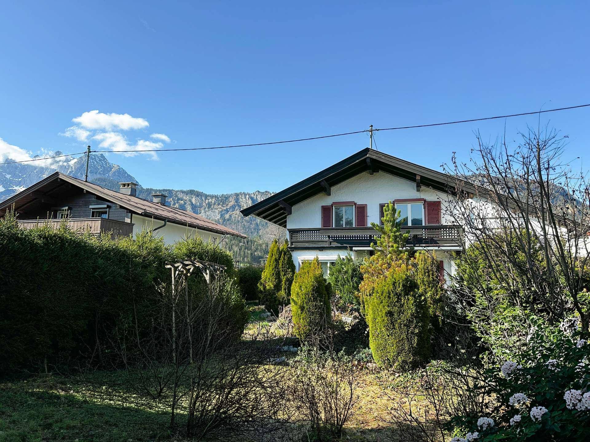 Traditionelles Haus mit Satteldach, Balkon und roten Fensterläden, umgeben von grüner Bepflanzung.