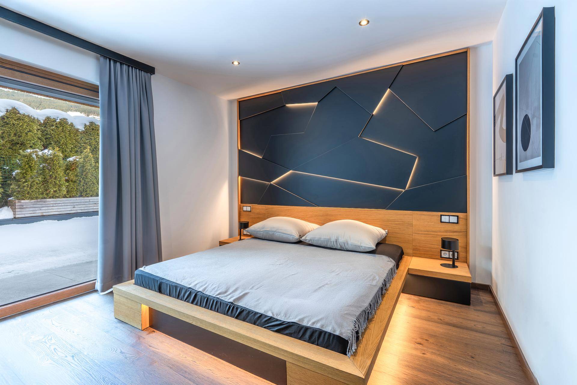 Stilvolles Schlafzimmer mit beleuchteter Akzentwand, Holzbett und großem Fenster mit Blick ins Grüne.