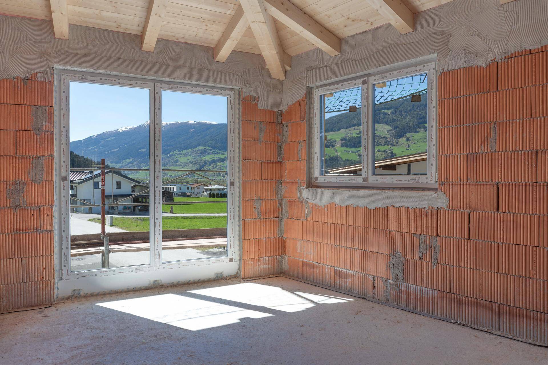 Rohbau eines Zimmers mit Ziegelwänden, neuen Fenstern und Blick auf die umliegende Berglandschaft.