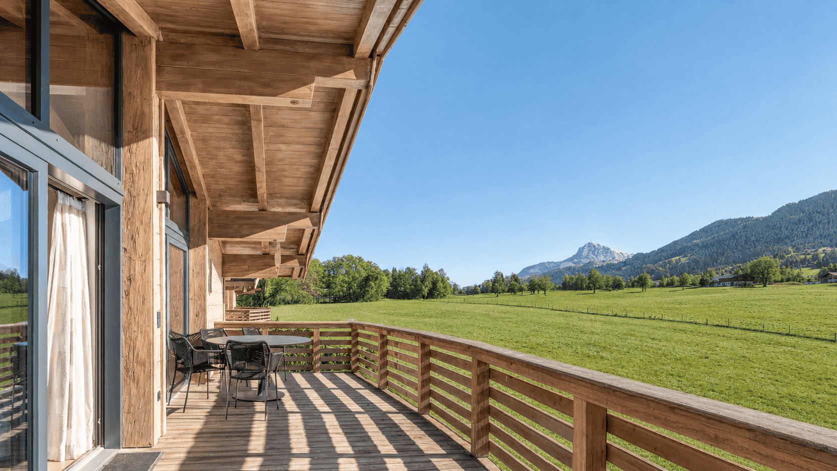Großer Holzbalkon mit Sitzgelegenheiten und weitem Blick auf grüne Wiesen und Berge.