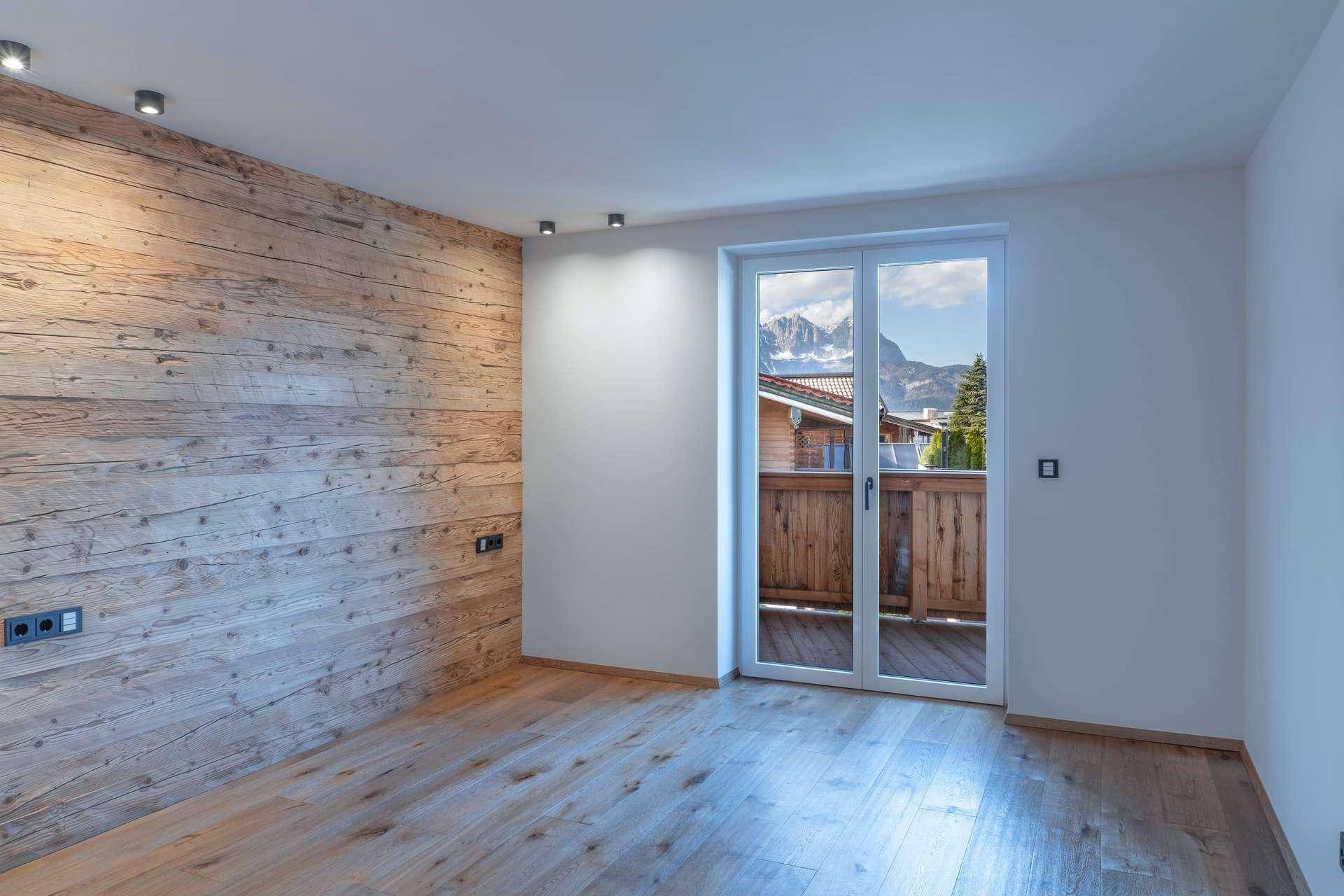 Heller Raum mit einer Holzakzentwand, Parkettboden und Balkontür mit Bergblick.