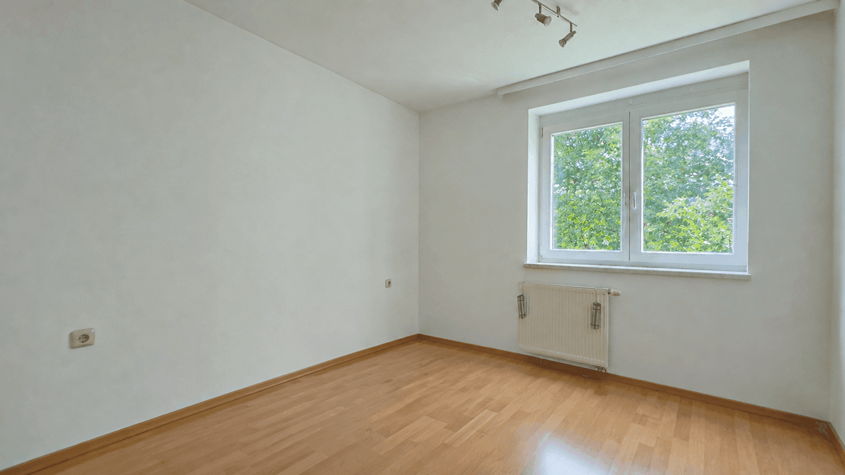 Freundliches Zimmer mit Parkettboden und großem Fenster mit Blick ins Grüne.
