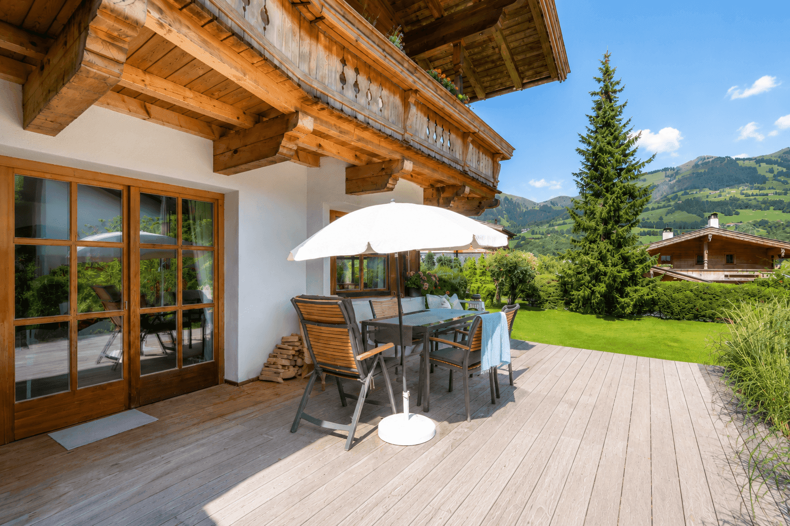 Sonnige Holzterrasse mit Esstisch, Sonnenschirm und Blick auf die umliegende Berglandschaft.