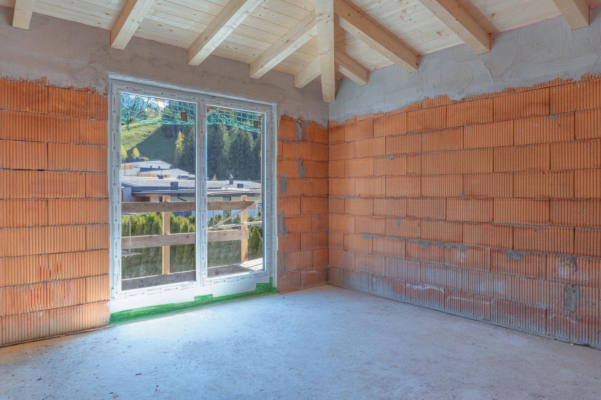 Rohbau eines Raumes mit Ziegelmauerwerk, Holzbalkendecke und großem Fenster mit Blick ins Grüne.