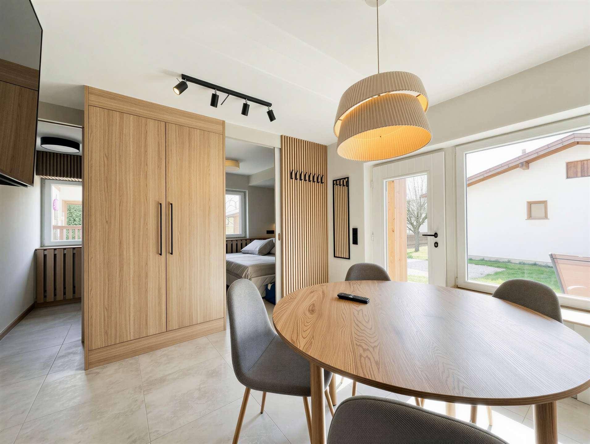 Offener Wohn- und Essbereich mit Holztisch, modernen Leuchten und Zugang zum Schlafzimmer.