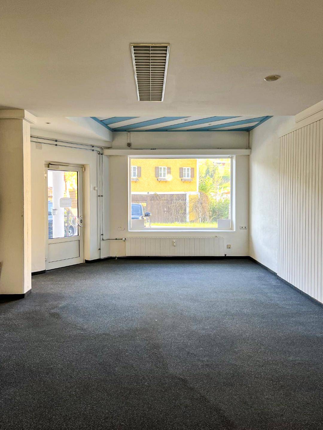 Leerer Gewerberaum mit Teppichboden, großer Fensterfront und Deckenlüftung, bereit zur Nutzung.