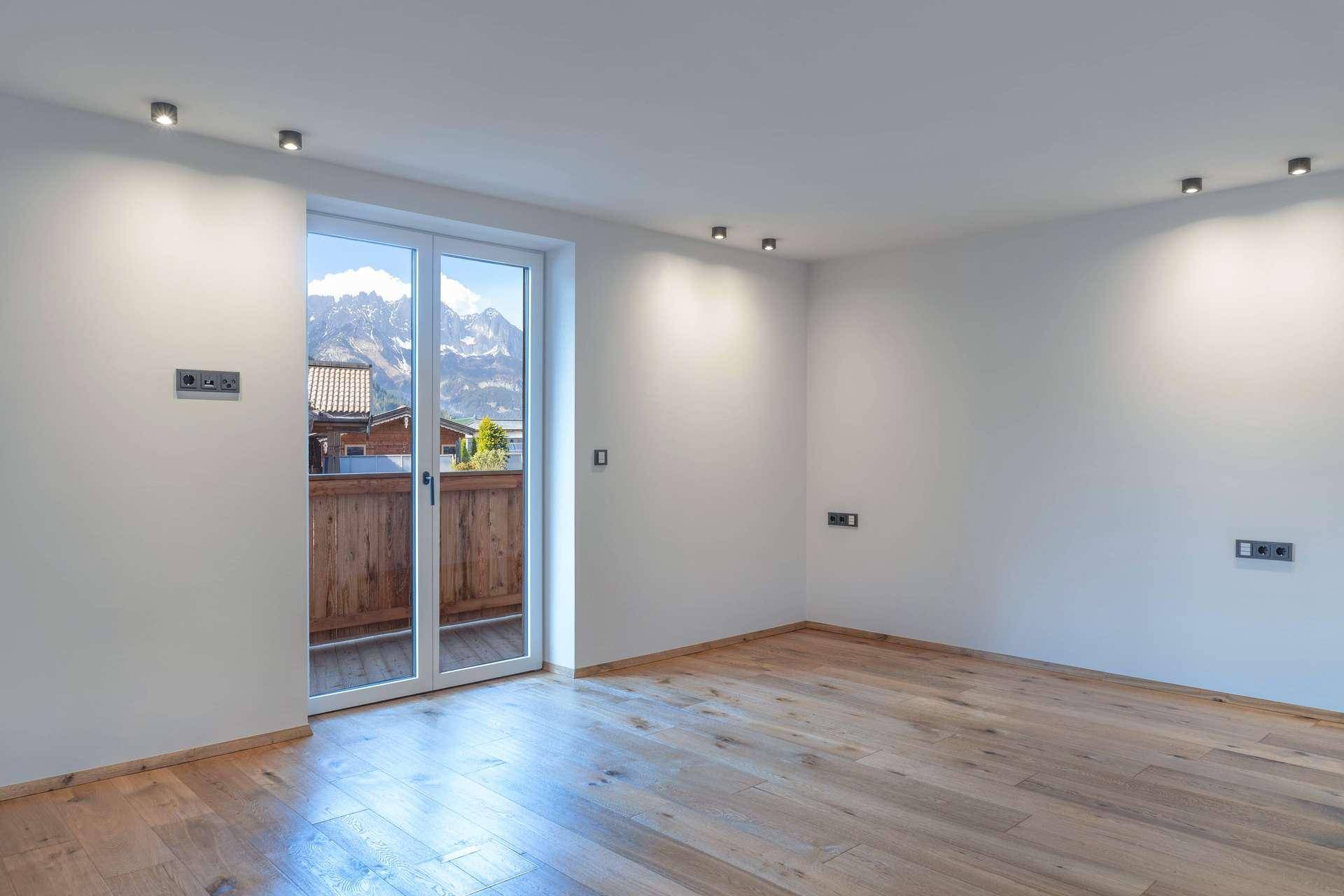Heller, leerer Raum mit Holzboden, modernen Deckenleuchten und Zugang zu einem Balkon.
