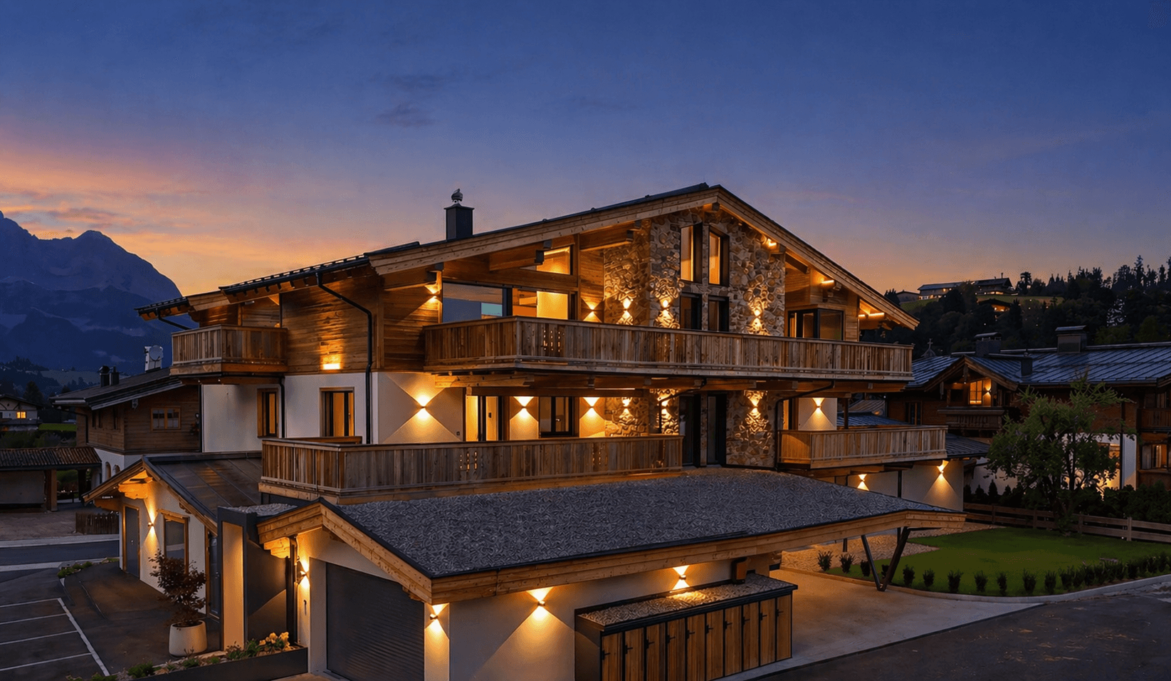 Beleuchtetes Chalet im alpinen Stil bei Dämmerung mit Garagen und Parkplätzen im Vordergrund.
