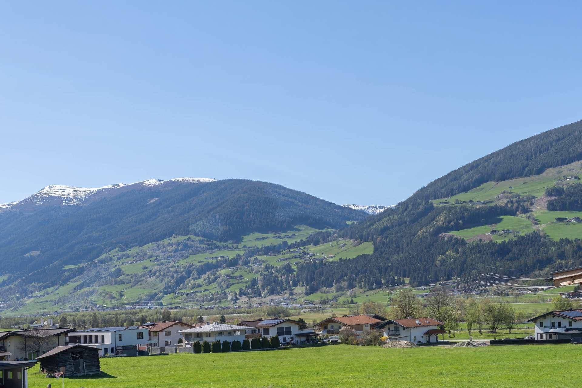 Weitläufige Landschaft mit grünen Hügeln, Bergen und einer Siedlung unter klarem blauem Himmel.