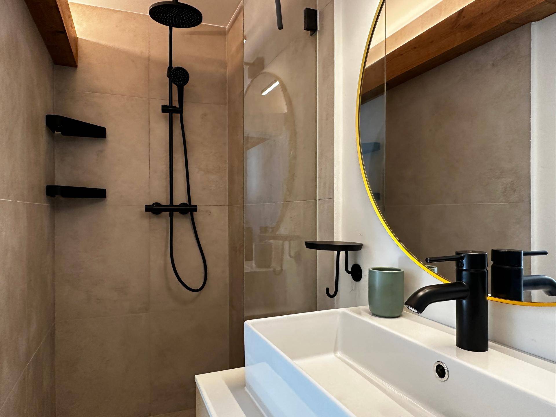 Modernes Badezimmer mit begehbarer Dusche, schwarzen Armaturen und einem runden Spiegel.