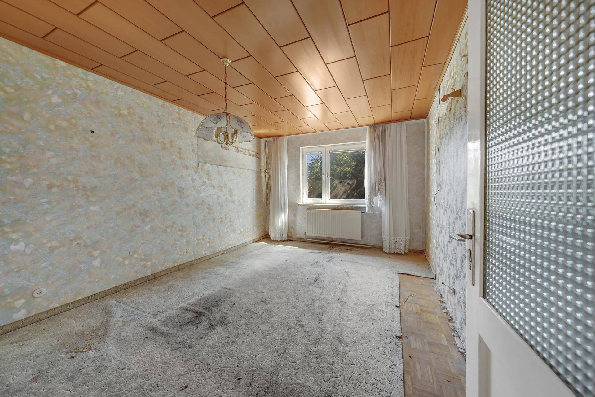 Schlafzimmer mit Teppichboden, Holzdecke und abgenutzten Tapeten, renovierungsbedürftig.