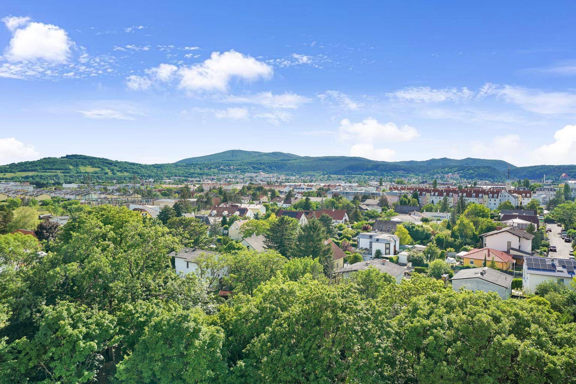 Weitläufiger Blick über die grüne Umgebung mit Wohngebäuden und Bergen im Hintergrund.