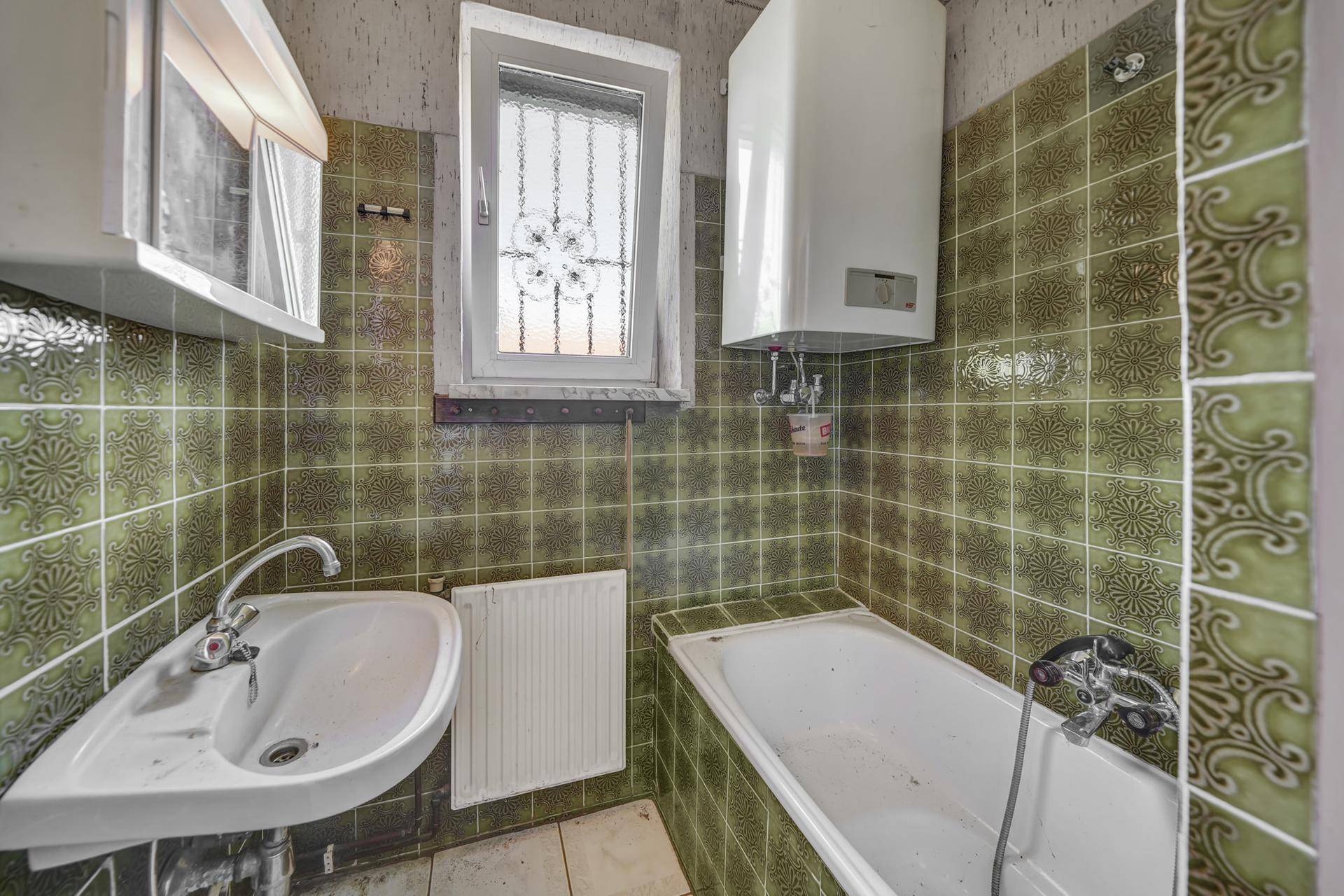 Badezimmer mit grünen Fliesen, Badewanne und kleinem Fenster, sehr renovierungsbedürftig.
