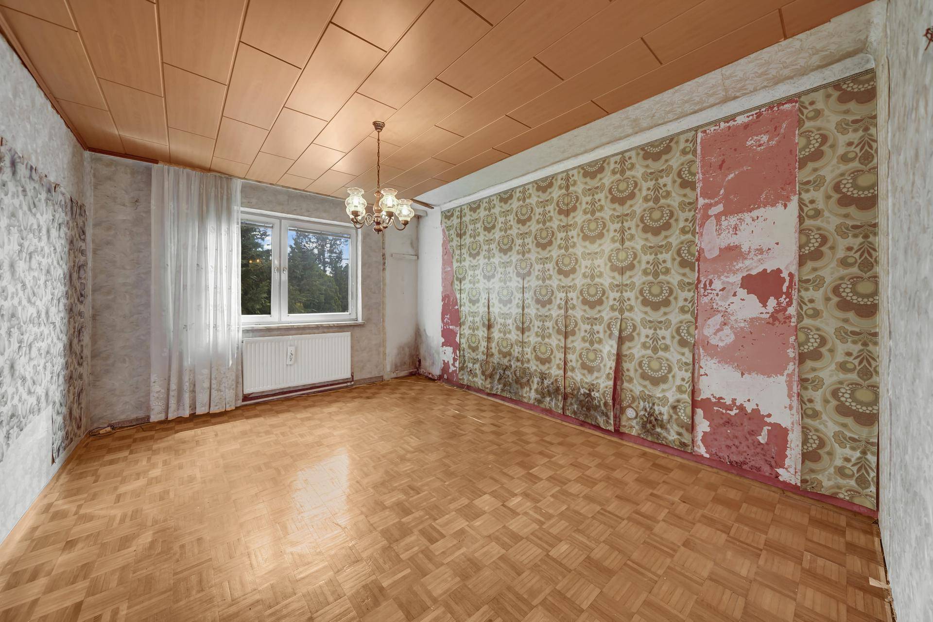 Geräumiges Zimmer mit Fischgrätparkett, Holzdecke und floraler Tapete, renovierungsbedürftig.
