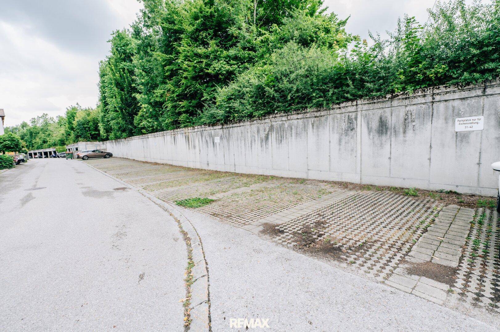 Außenparkplätze mit Betonmauer im Hintergrund und umgebenden Bäumen.