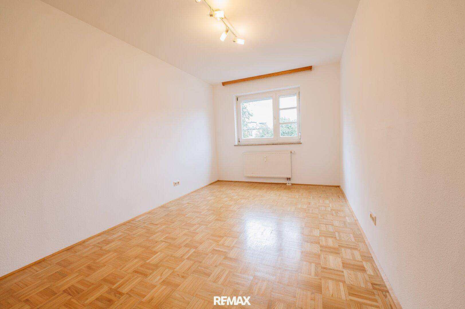 Geräumiges Zimmer mit hellem Parkettboden und einem großen Fenster, das viel Tageslicht hereinlässt.