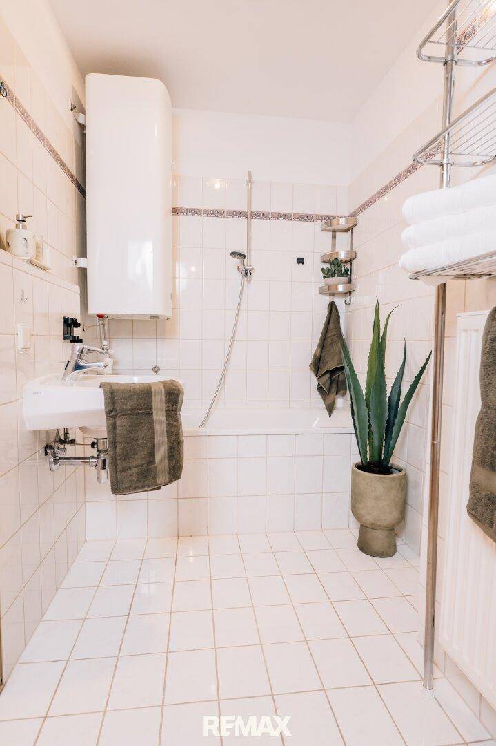 Badezimmer mit weißen Fliesen, Badewanne, Waschbecken und einem Warmwasserboiler.
