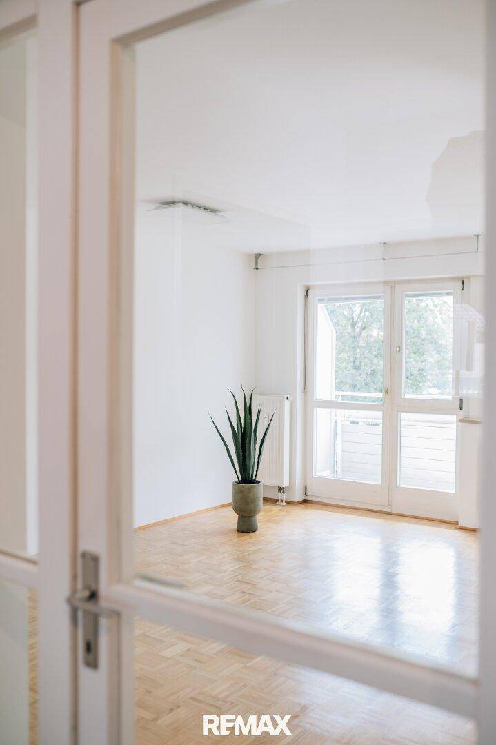 Blick durch eine Glastür in ein helles Zimmer mit Parkettboden und Zugang zum Balkon.