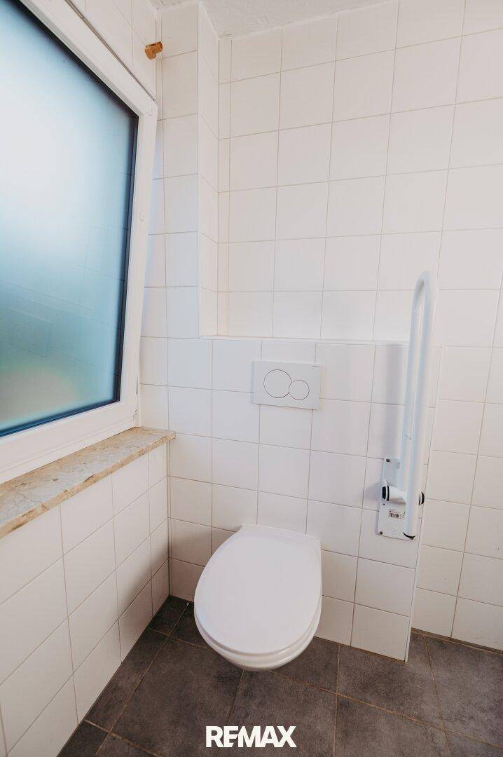 Kleines WC mit weißen Fliesen, Wand-WC und Haltegriffen für Barrierefreiheit.