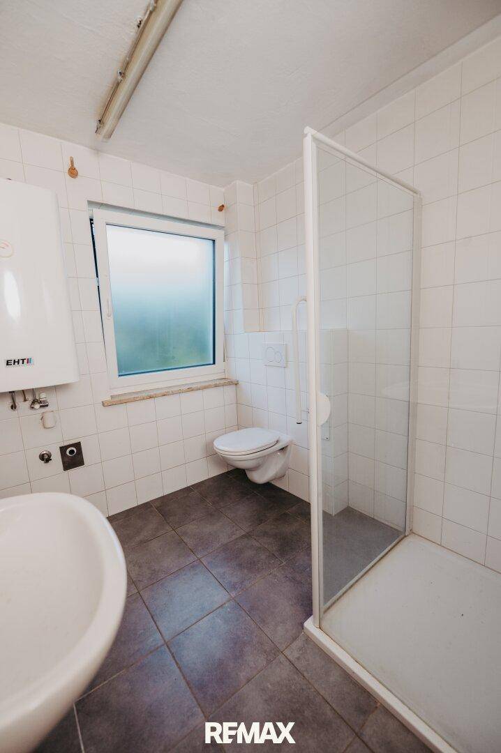 Badezimmer mit Dusche, WC, Waschbecken und Fenster, das eine Renovierung benötigt.