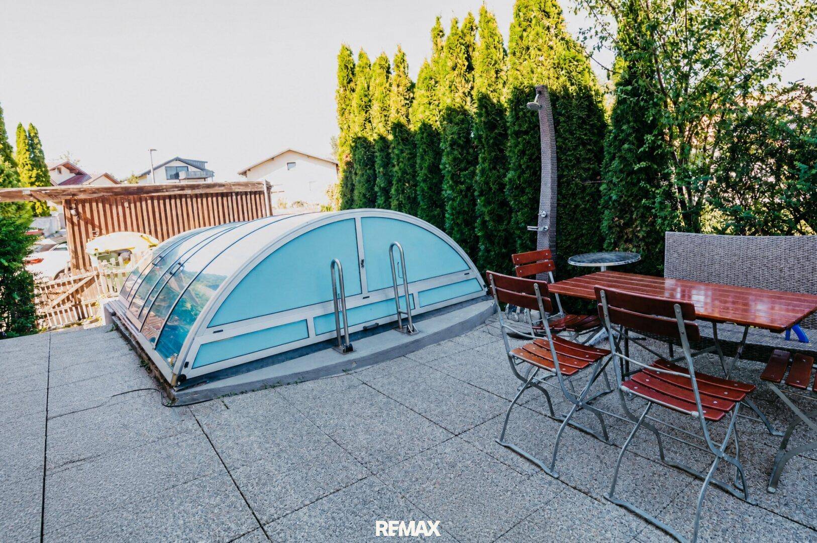 Geflieste Terrasse mit Gartenmöbeln und überdachtem Pool, umgeben von hohen Hecken.