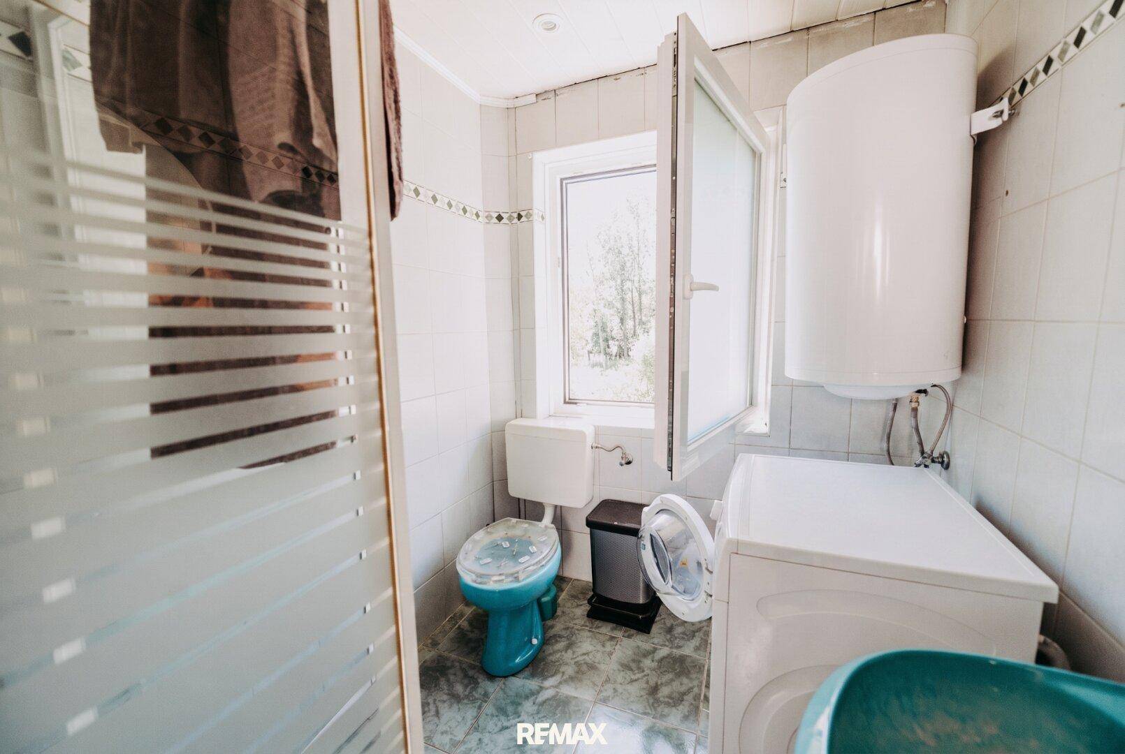 Badezimmer mit Dusche, WC, Waschmaschine und Fenster, das eine Modernisierung benötigt.
