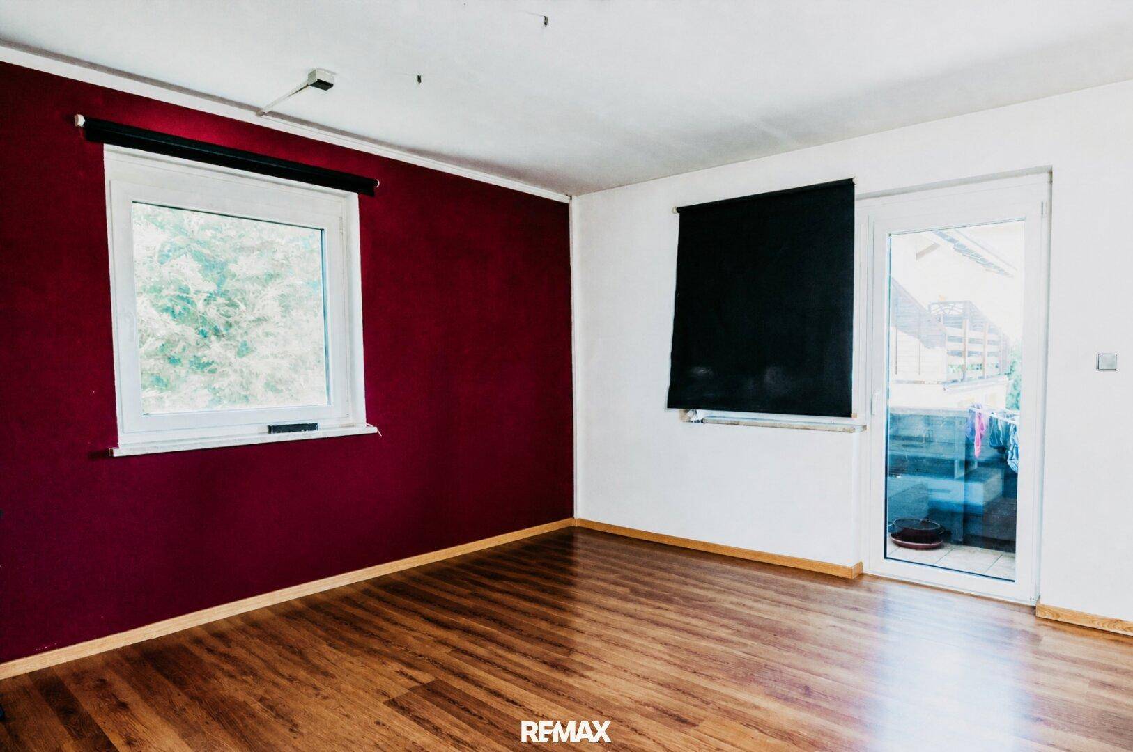 Heller Raum mit Holzboden, roter Akzentwand und Zugang zu einem Balkon oder einer Terrasse.