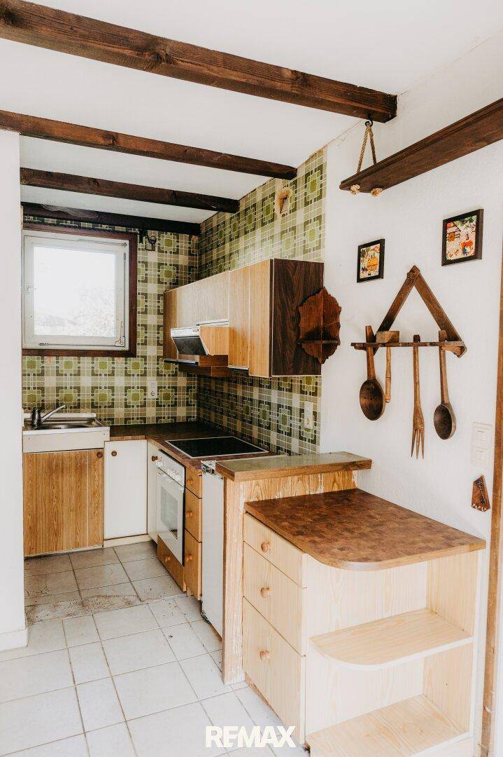 Rustikale Küche mit älteren Holzschränken, gemusterter Tapete und sichtbaren Holzbalken.