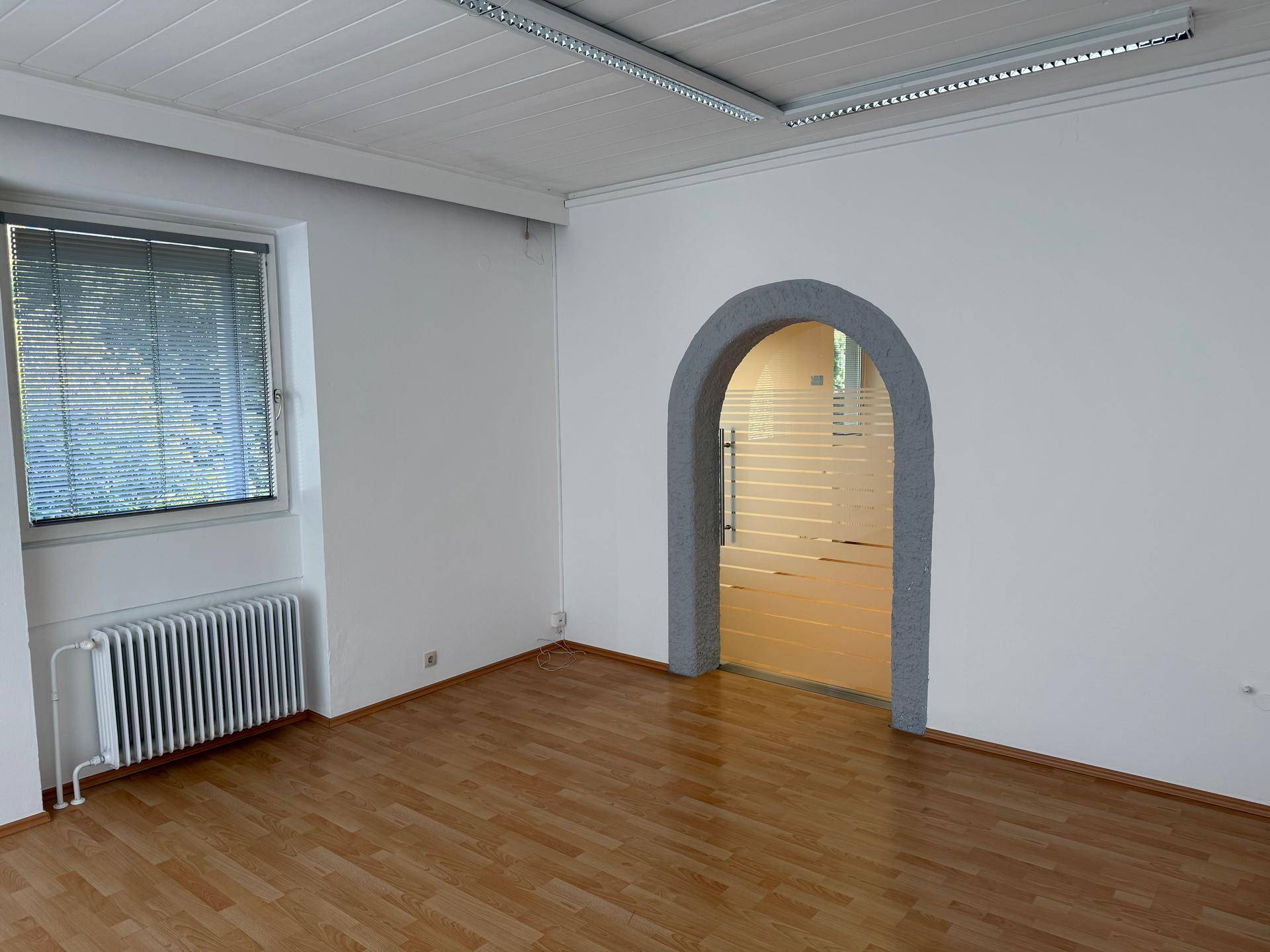 Heller Büroraum mit Laminatboden, einem Fenster mit Jalousien und einem eleganten Rundbogen zu einem weiteren Bereich.
