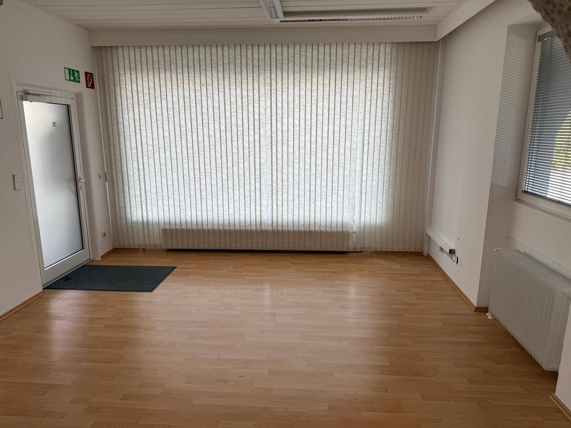 Großer, heller Büroraum mit Laminatboden, einer breiten Fensterfront mit Vertikaljalousien und einem Ausgang.