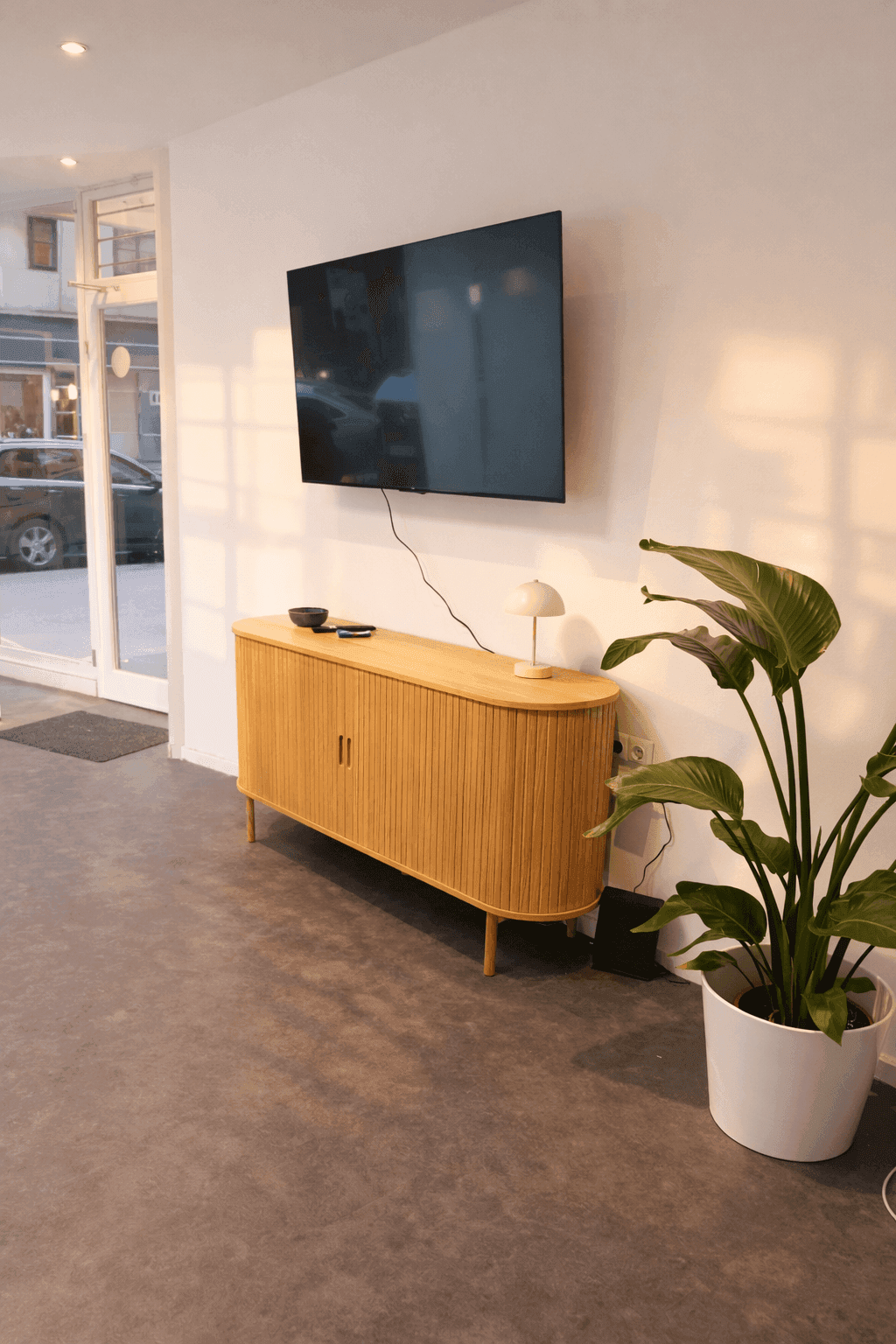 Heller Bürobereich mit einem modernen Sideboard, Wandfernseher und einer großen Pflanze.